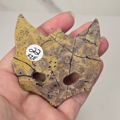 A hand holding a small cat-shaped decorative piece made from a mottled yellow and brown stone with black veining and two circular cutouts. A small white sticker with handwritten text is attached near one ear.