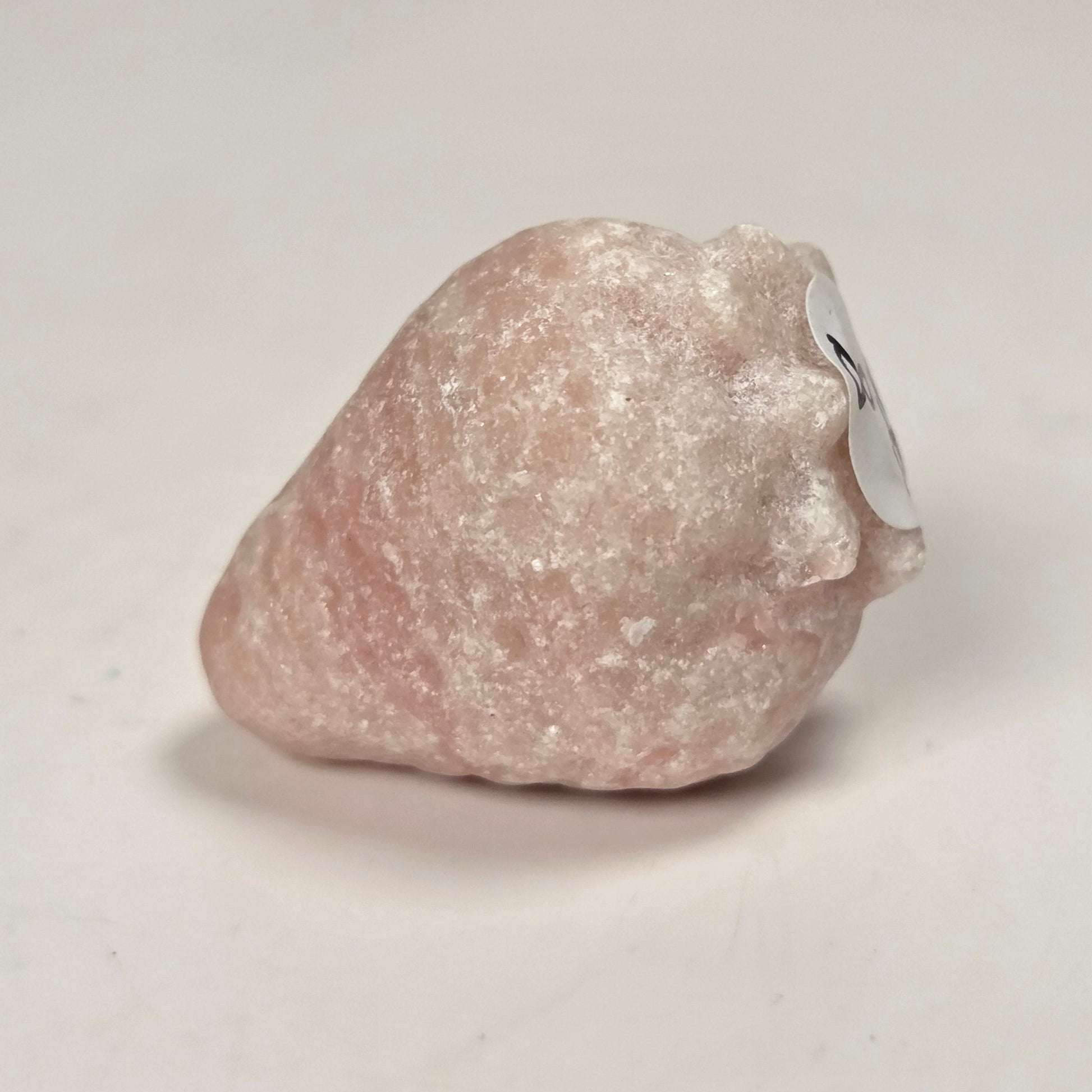 A small, rough, pale pink stone or crystal with a small white tag attached, photographed on a plain light background.
