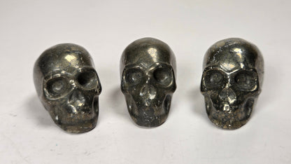 Three small metallic skull-shaped objects arranged side by side on a plain light background.