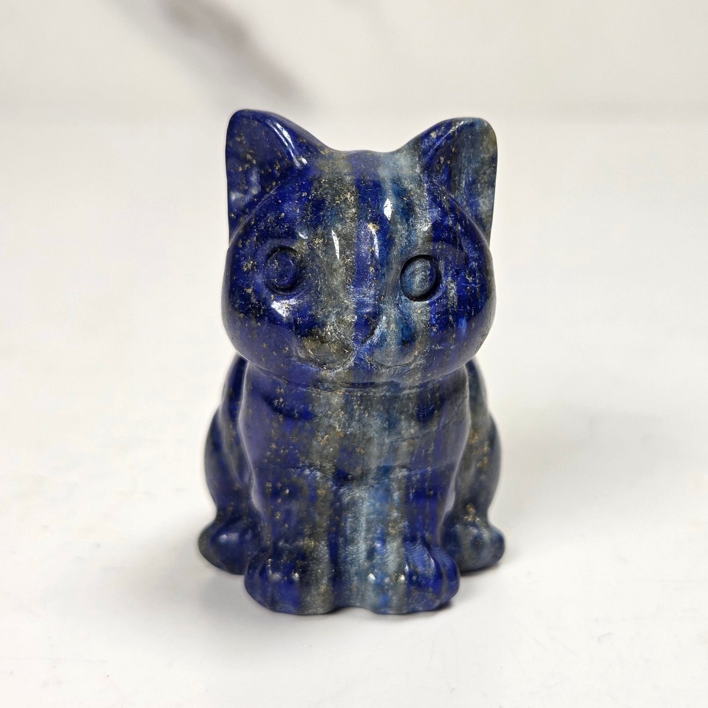 A small carved cat figurine made of polished lapis lazuli, featuring deep blue and black marbling with subtle golden flecks, set against a light off-white background.