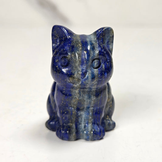 A small carved cat figurine made of polished lapis lazuli, featuring deep blue and black marbling with subtle golden flecks, set against a light off-white background.