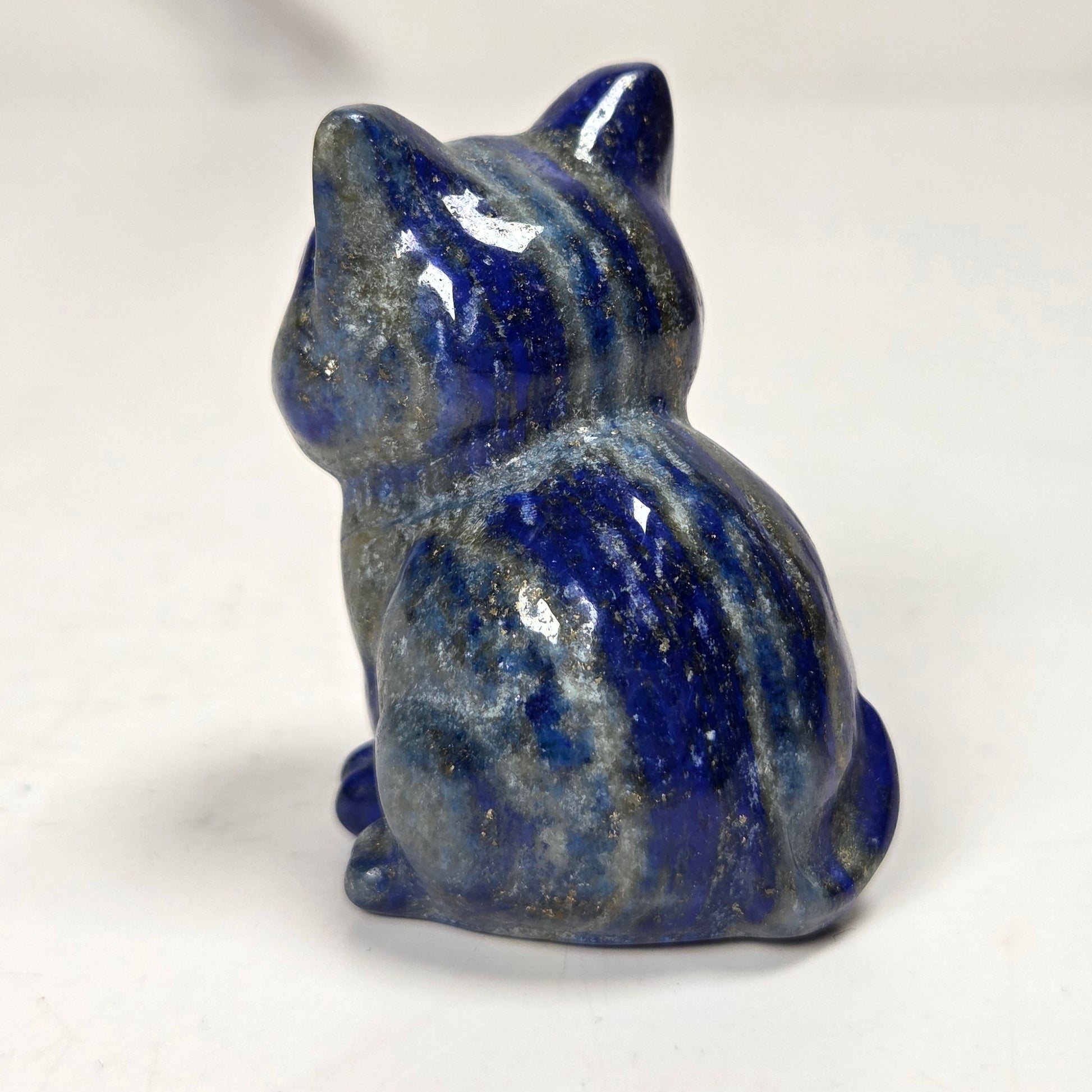 A small glossy blue cat figurine with natural stone speckling, shown from the back on a light gray surface.