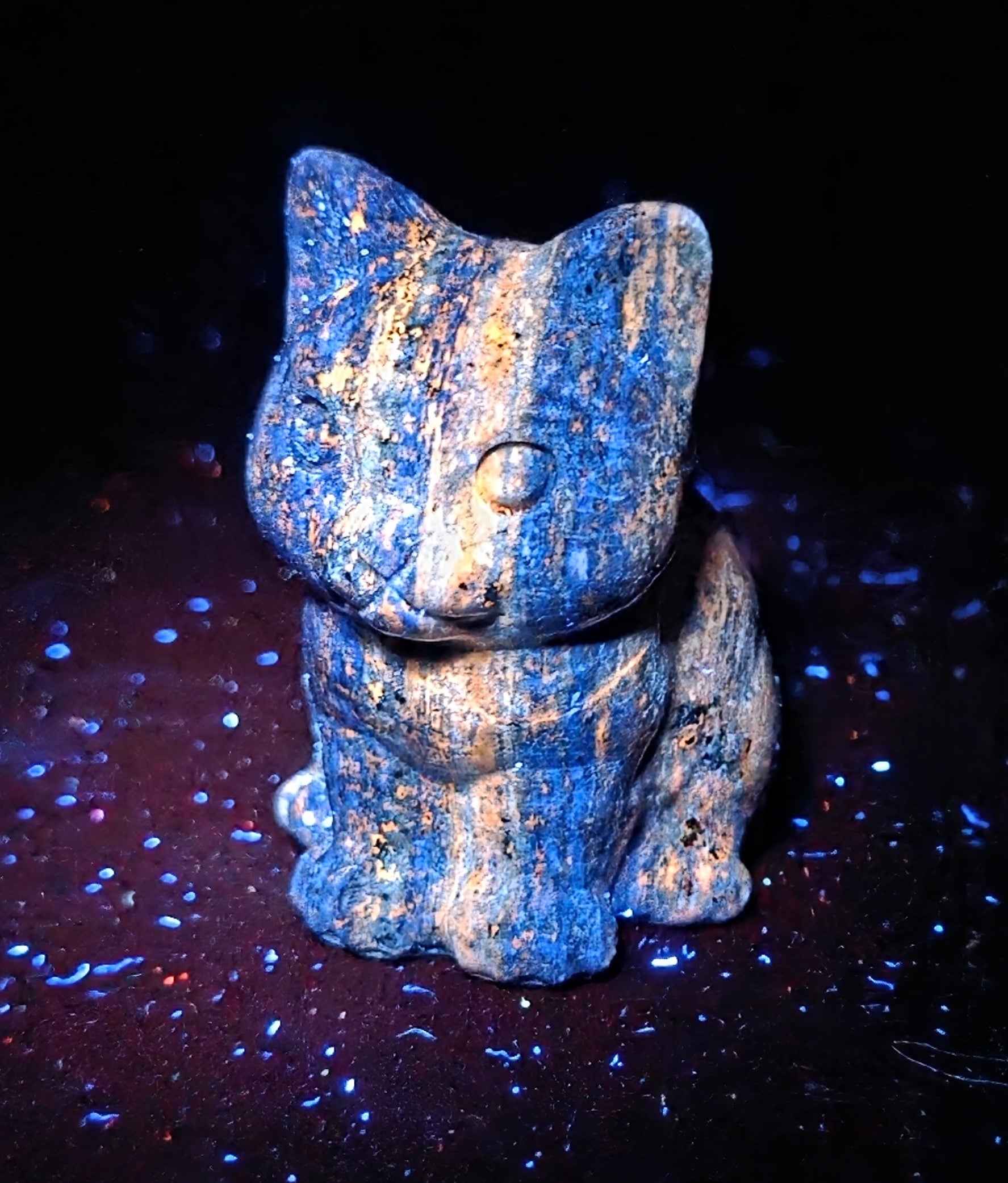 A small blue and tan cat figurine, likely made of stone or ceramic, standing on a dark surface with blue speckles against a black background.