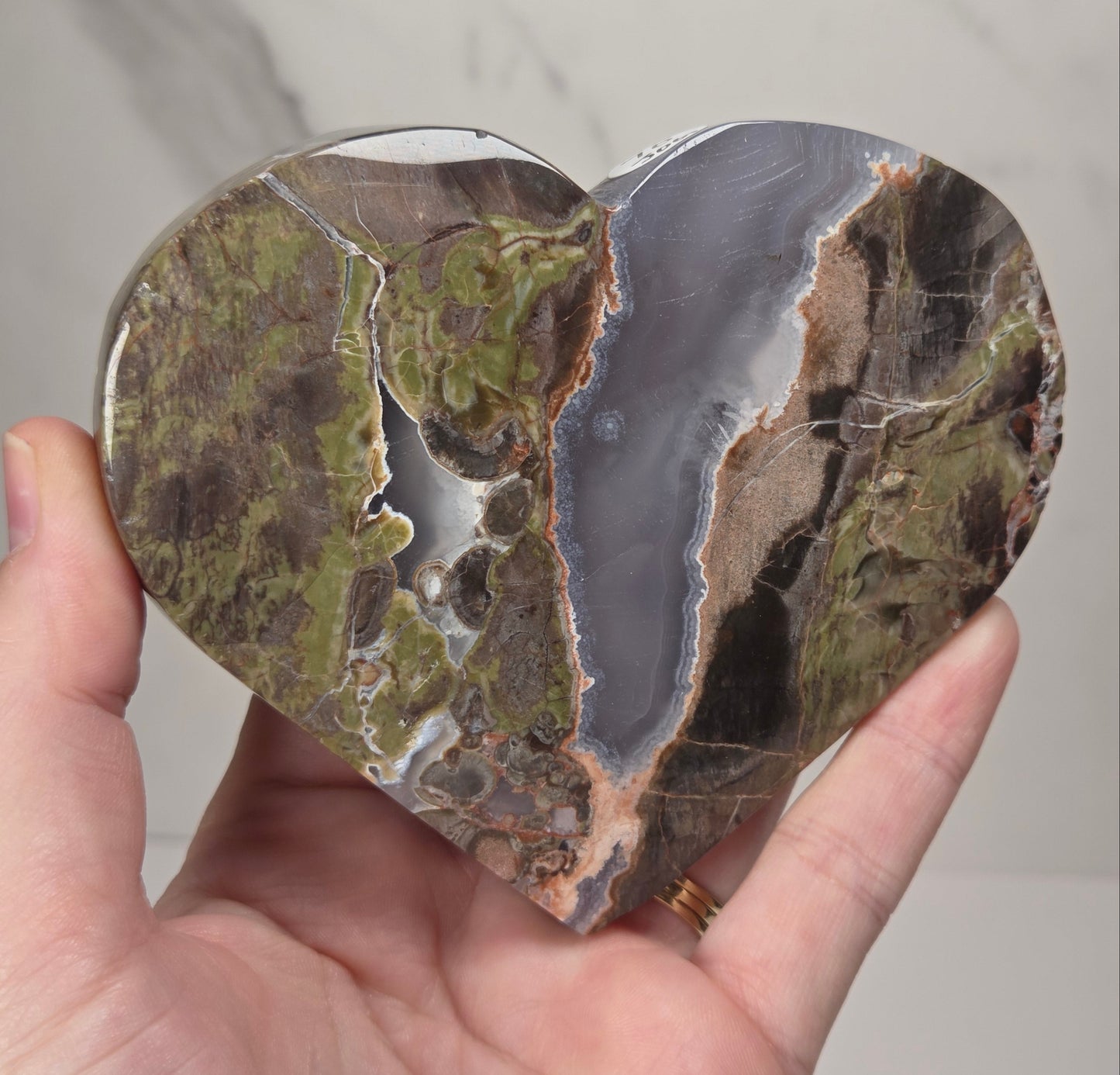 A heart-shaped polished stone sculpture with natural earthy patterns in olive green, brown, gray, and beige, held in a person's hand against a light marble background.