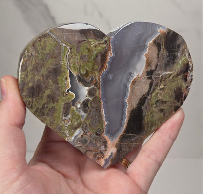 A heart-shaped polished stone sculpture with natural earthy patterns in olive green, brown, gray, and beige, held in a person's hand against a light marble background.