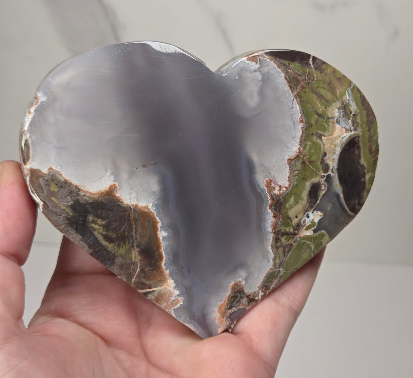 A heart-shaped polished agate stone held in a person's hand, showing natural banding with gray, white, green, and brown tones against a light marble-like background.