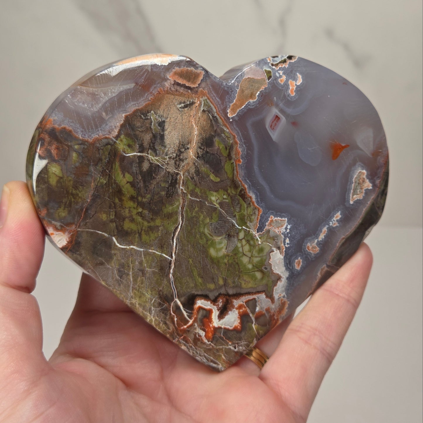 A heart-shaped polished agate stone with natural earthy patterns, held in a person's hand against a light gray marble-patterned background.