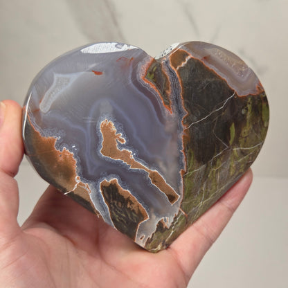 A polished heart-shaped agate stone with natural banding in gray, white, brown, and green, held in a person's hand against a light marble-patterned background.