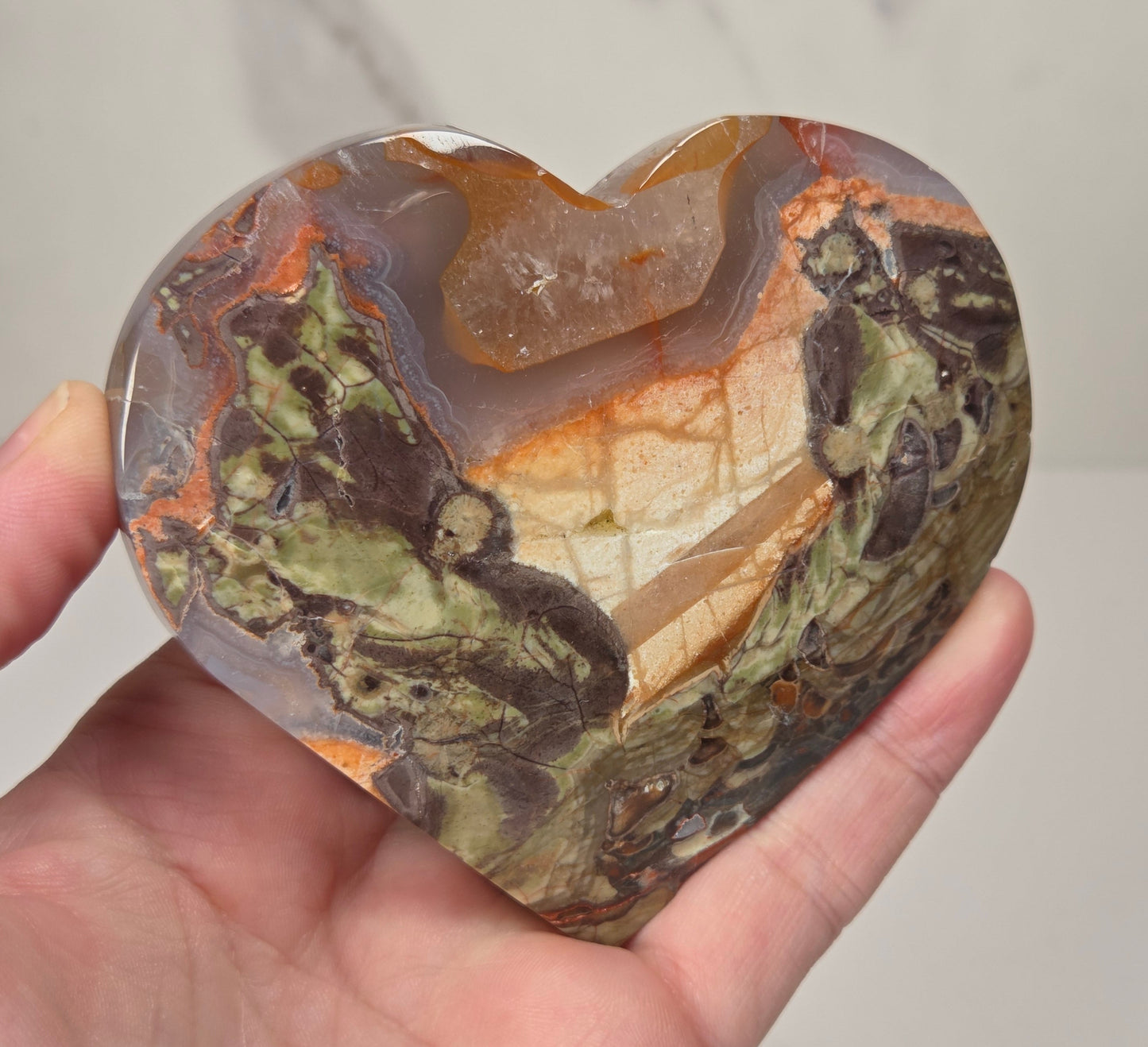 A heart-shaped polished agate crystal holding a small quartz point, held in a person's hand against a light marble background.