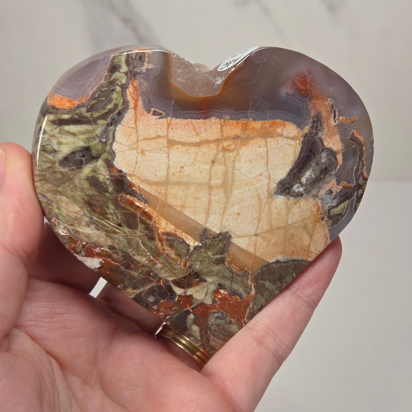 A heart-shaped polished agate stone with natural earthy patterns, held in a person's hand against a light gray marble-patterned background.