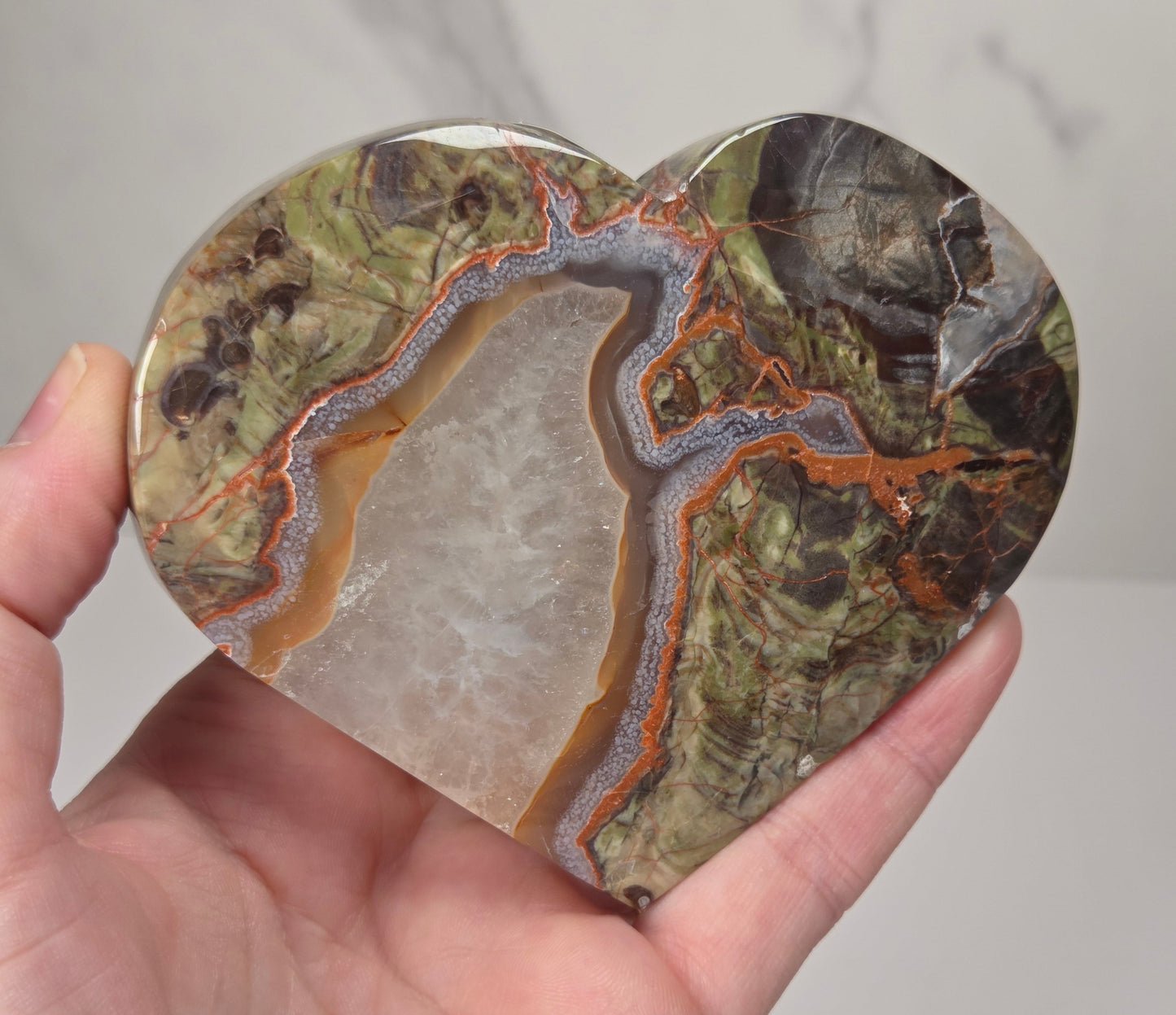 A heart-shaped polished agate stone featuring a natural geode pattern with bands of green, brown, orange, and a translucent white center, held in a person's hand against a light gray marble background.