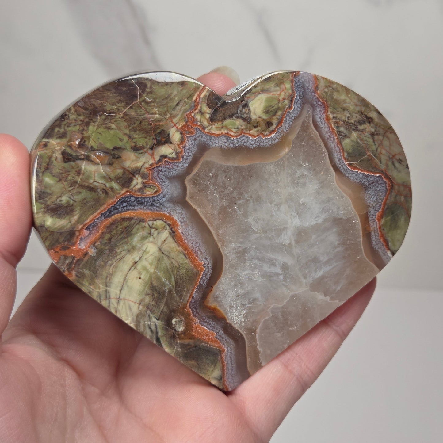 A heart-shaped polished stone or agate slab with natural marbled patterns in olive green, gray, brown, and orange, held in a person's hand against a light marble-patterned background.
