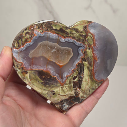A heart-shaped polished agate stone with a natural cross-section, held in a person's hand against a light marble-patterned background.