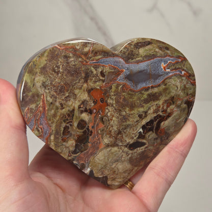 A heart-shaped polished stone with natural marbled patterns in brown, green, orange, and blue-gray, held in a person's hand against a light marble-patterned background.