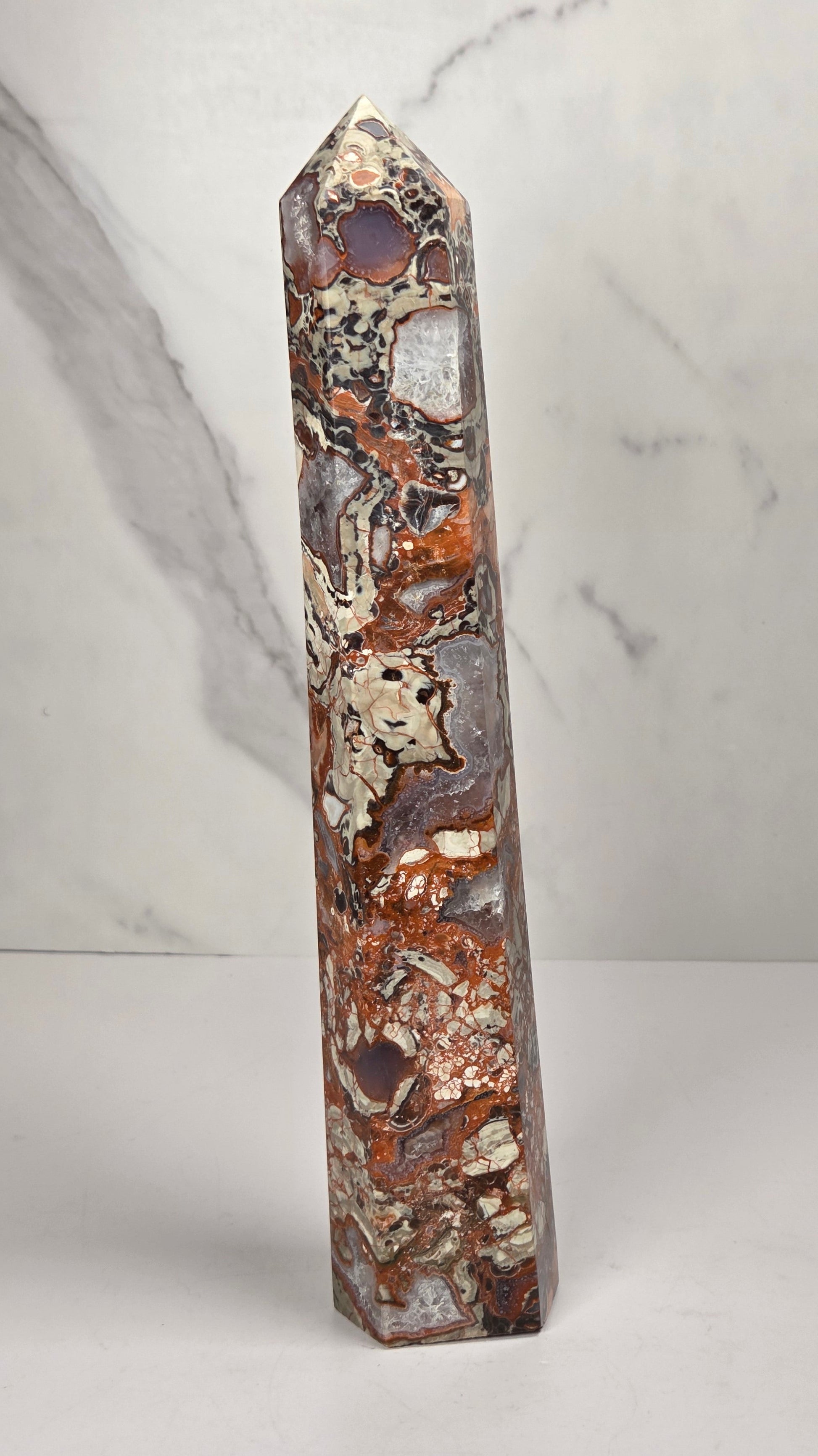 A tall, narrow decorative crystal tower made of natural copper pyrite and silver quartz with brown and orange mineral inclusions, standing upright on a white surface against a light gray marble-patterned background.