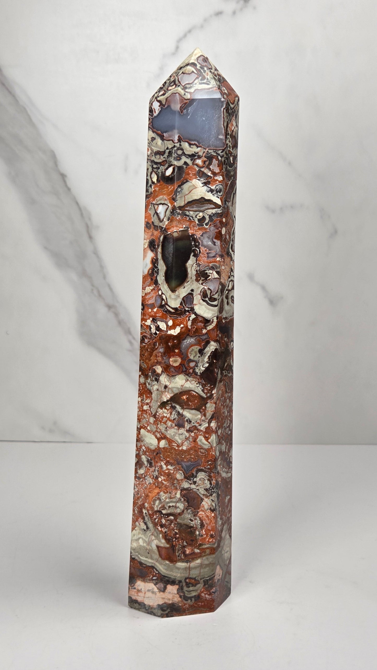 A polished natural mineral obelisk featuring a marbled pattern of rust-red, brown, cream, and black, standing upright on a white surface against a light gray marble-patterned background.