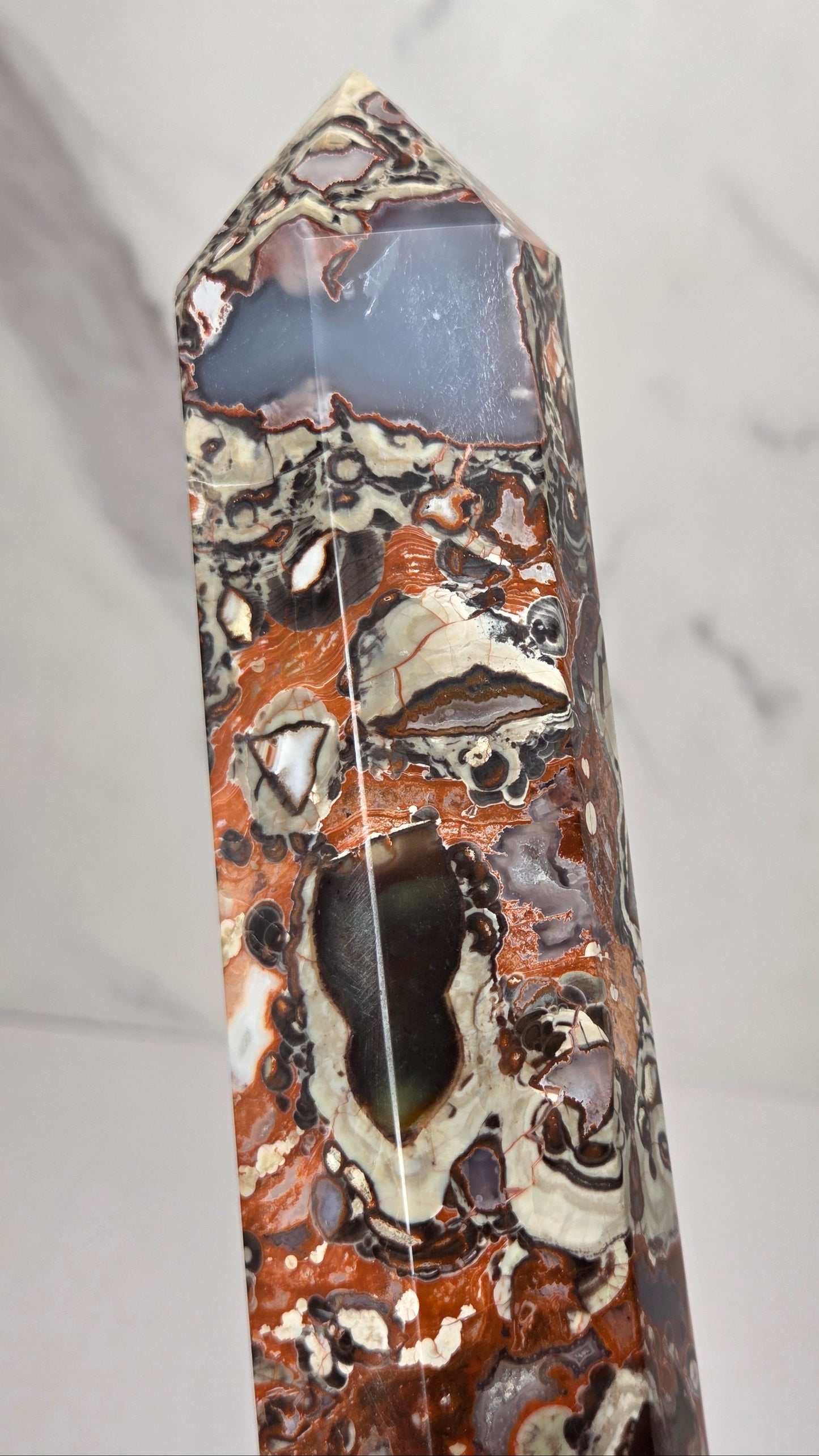 A polished natural crystal point featuring a marbled pattern of rust orange, cream, brown, and dark gray, standing upright against a light gray marble-patterned background.