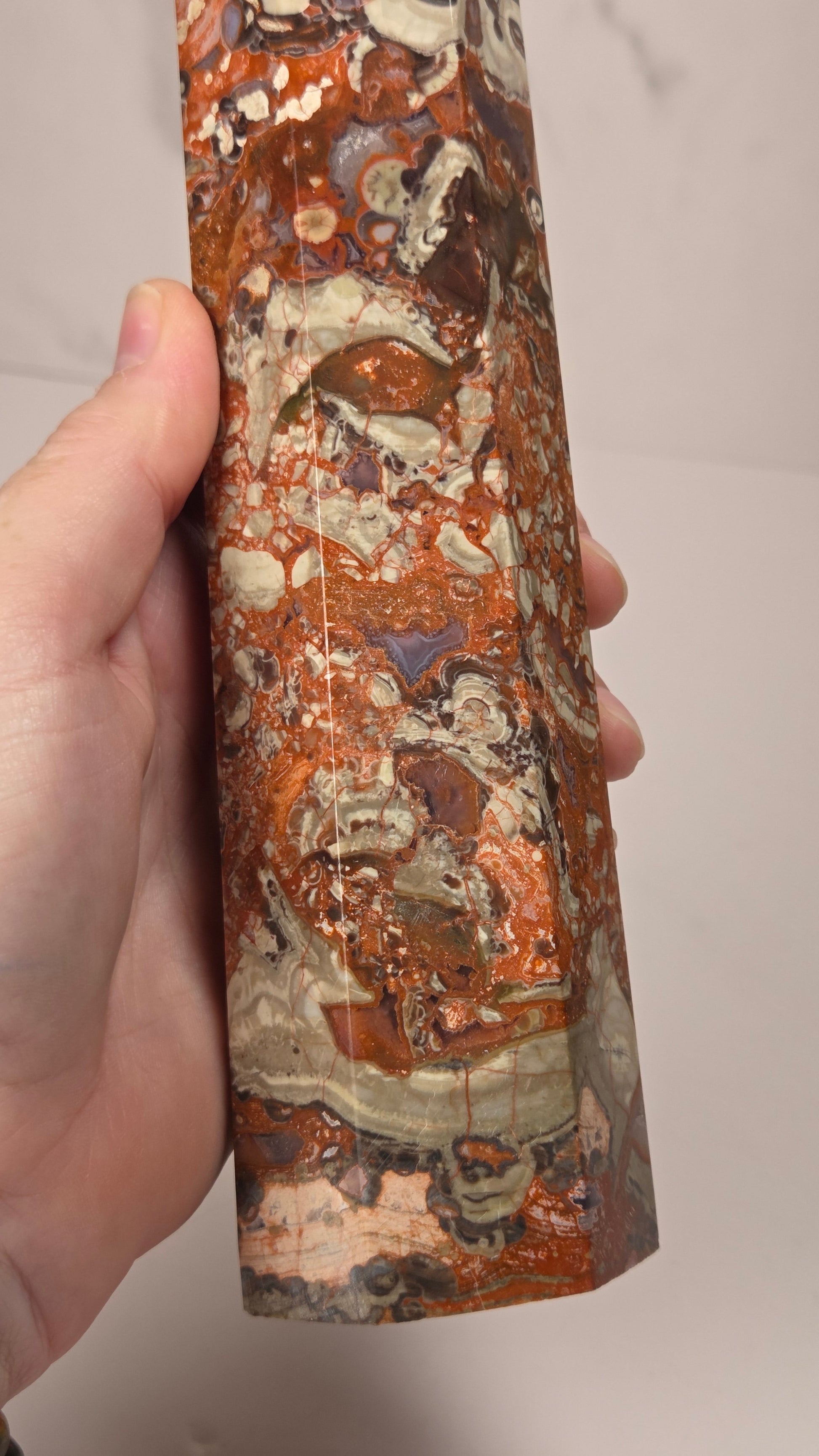 A hand holding a polished cylindrical stone specimen showing orange, brown, cream, and gray marbled patterns.