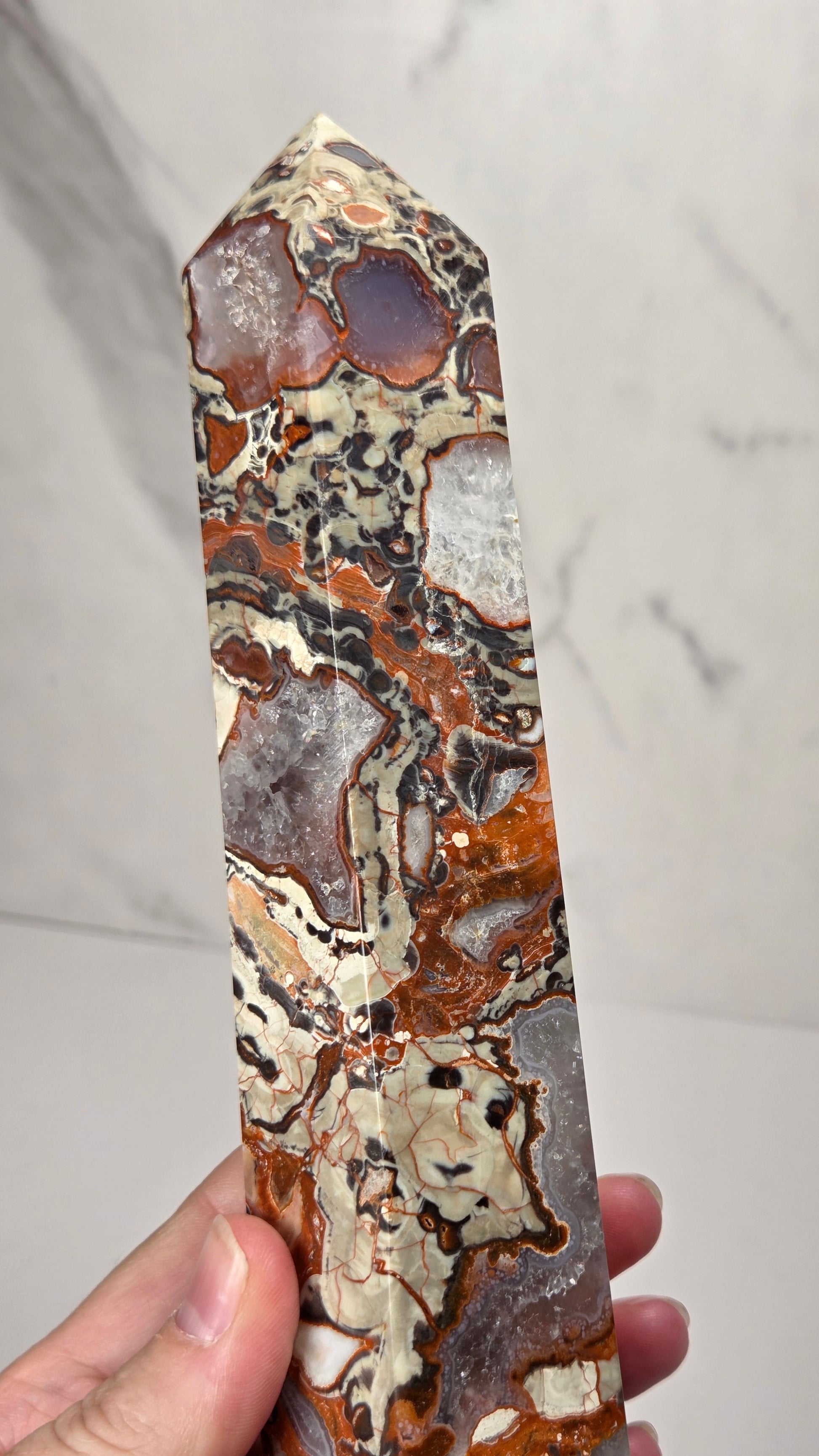 A hand holds a polished natural crystal point showing intricate patterns of brown, beige, gray, and black agate and jasper.