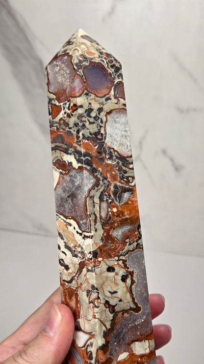 A hand holds a polished natural crystal point showing intricate patterns of brown, beige, gray, and black agate and jasper.