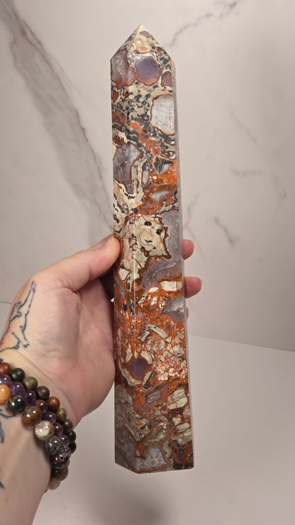 A hand with tattoos holds a polished natural quartz crystal obelisk showing orange, brown, and gray agate patterns, against a light gray marble-patterned background.