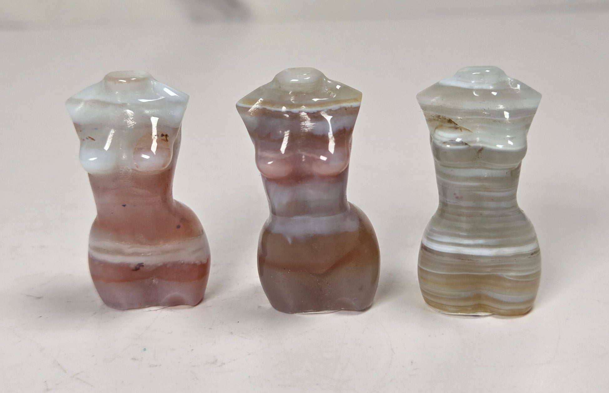 Three small carved agate figurines shaped like stylized female torso forms, displayed side by side on a light surface. The left figure is white and dusty rose, the center is brown and mauve, and the right is beige and cream with horizontal banding.