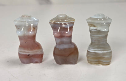 Three small carved agate figurines shaped like stylized female torso forms, displayed side by side on a light surface. The left figure is white and dusty rose, the center is brown and mauve, and the right is beige and cream with horizontal banding.