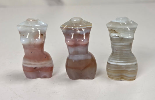 Three small carved agate figurines shaped like stylized female torso forms, displayed side by side on a light surface. The left figure is white and dusty rose, the center is brown and mauve, and the right is beige and cream with horizontal banding.