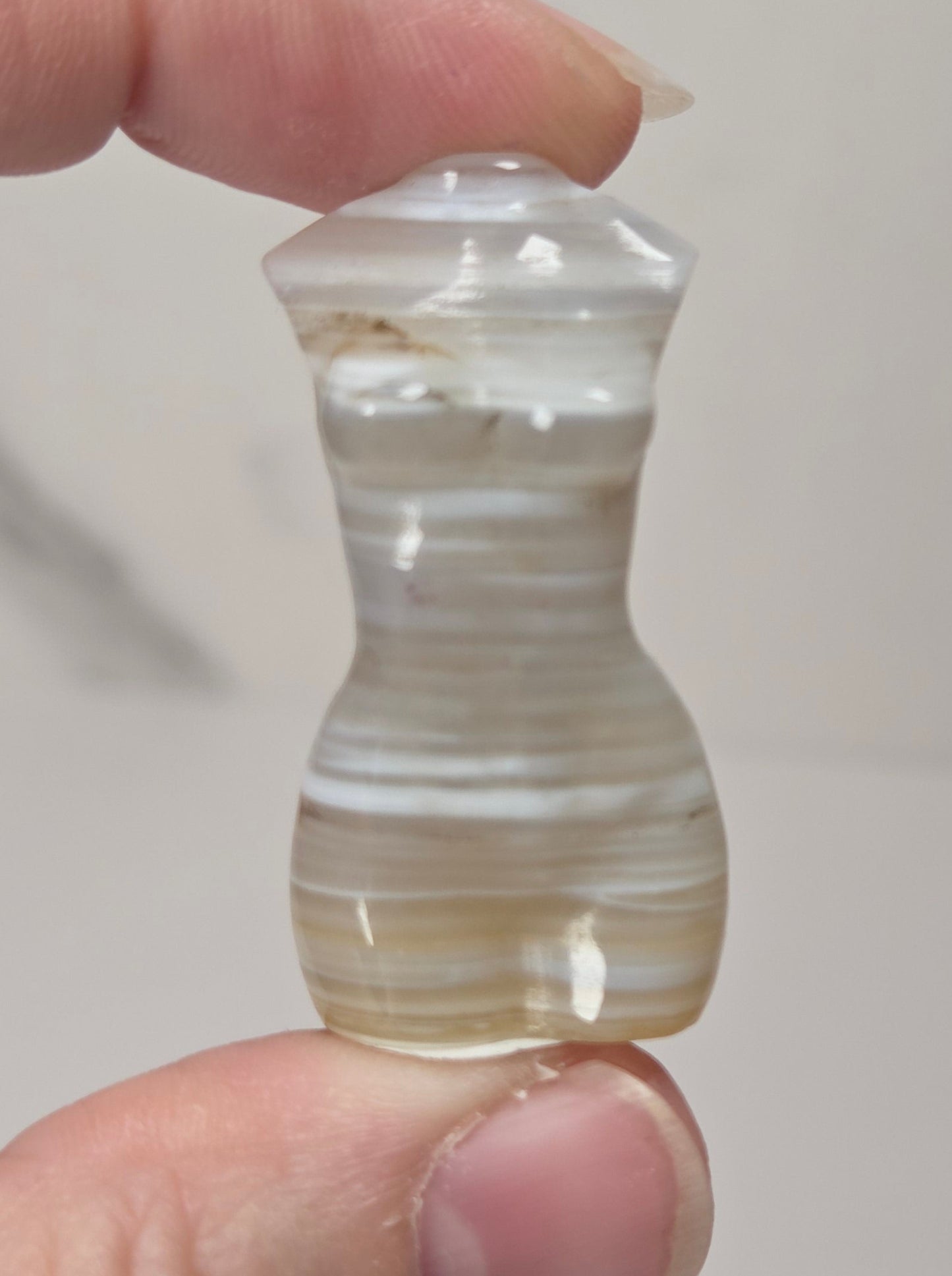 A hand holding a small, hourglass-shaped carved stone bead or pendant with horizontal banded patterns in cream, white, and light brown, set against a neutral light gray background.