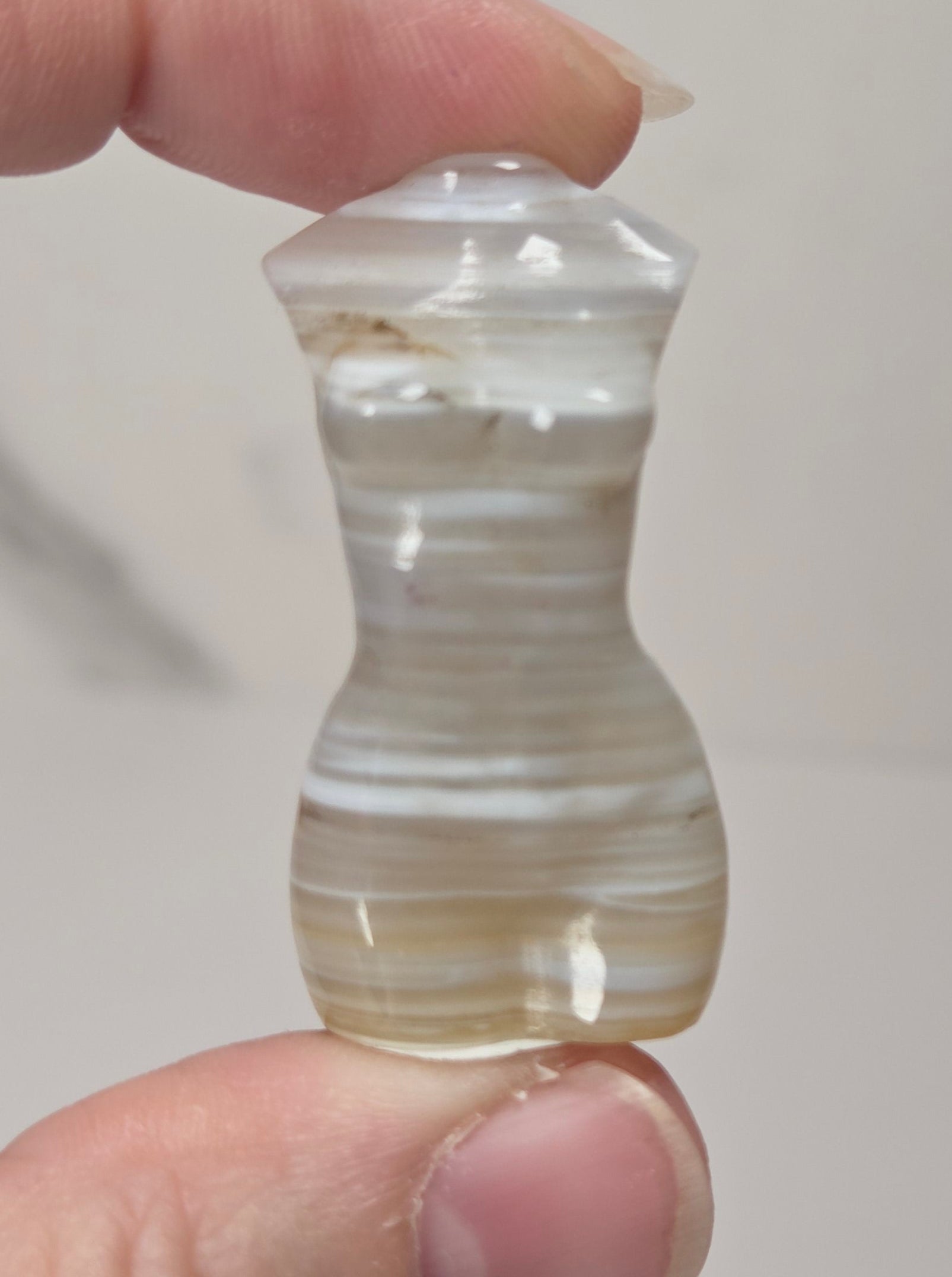 A hand holding a small, hourglass-shaped carved stone bead or pendant with horizontal banded patterns in cream, white, and light brown, set against a neutral light gray background.