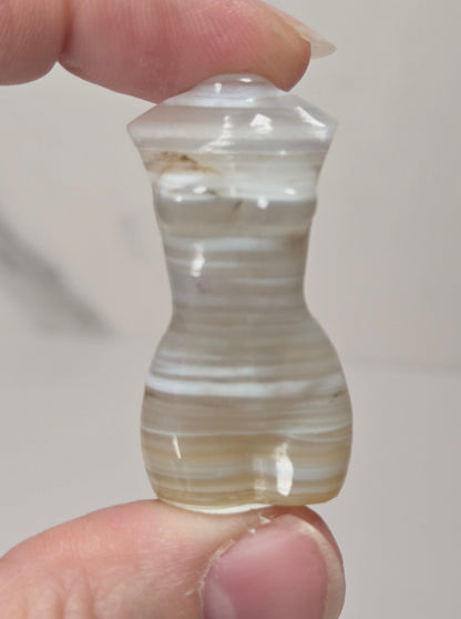 A hand holding a small, hourglass-shaped carved stone bead or pendant with horizontal banded patterns in cream, white, and light brown, set against a neutral light gray background.