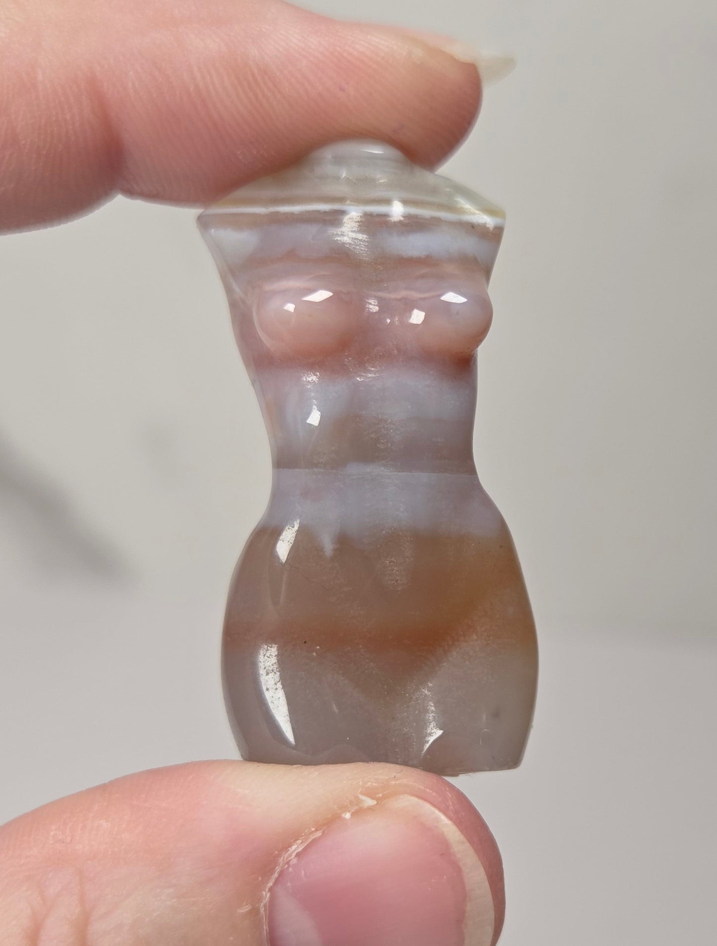 A small carved stone figurine of a female torso, held between two fingers to show its size. The figurine is made of a polished agate-like stone with horizontal bands of translucent white, light gray, and warm brown.