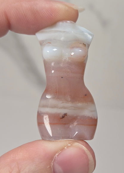 A hand holding a small carved stone figurine shaped like a torso, made from a translucent pinkish stone with white bands and small dark inclusions.