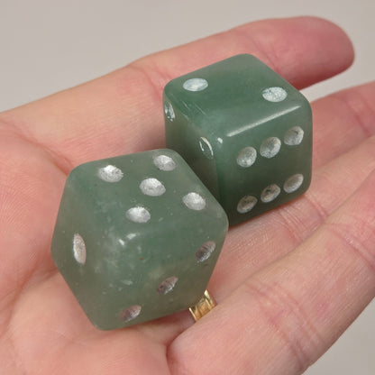Two translucent green dice-shaped jewelry pieces resting on an open hand, with a white marble-patterned background.