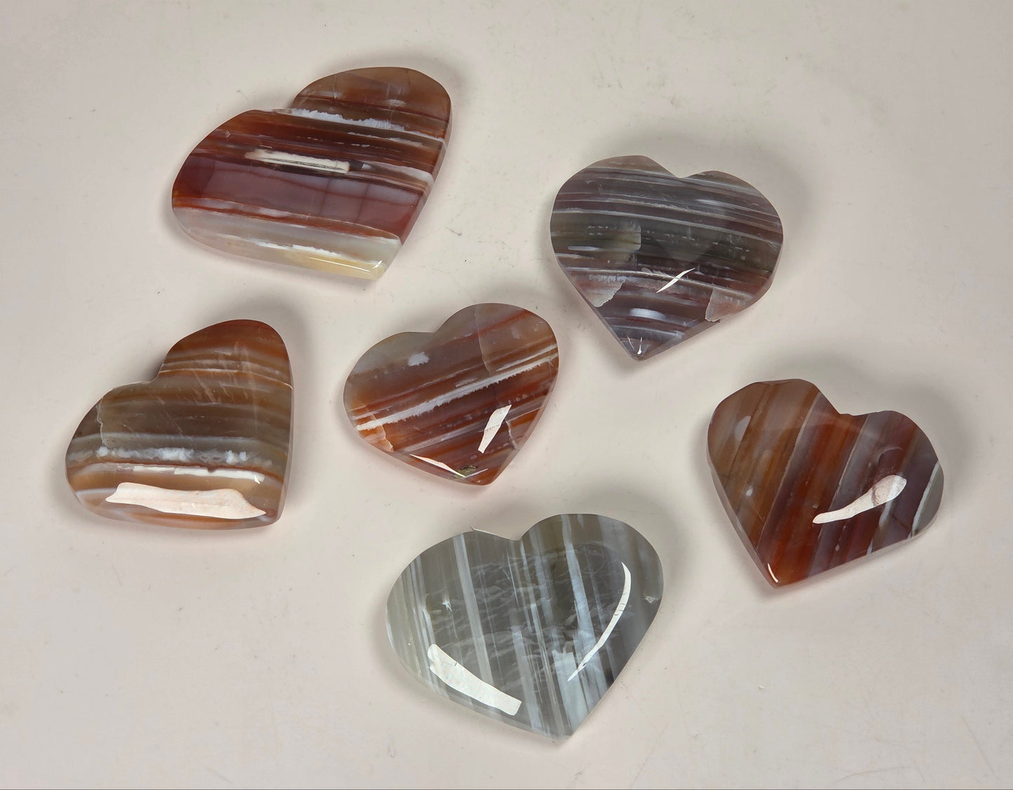 Six polished heart-shaped agate stones with natural banding patterns in shades of brown, gray, white, and reddish tones, arranged on a light beige surface.