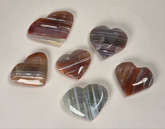 Six polished heart-shaped agate stones with natural banding patterns in shades of brown, gray, white, and reddish tones, arranged on a light beige surface.