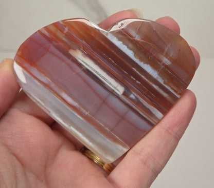A heart-shaped polished agate stone with horizontal bands of brown, orange, and white, held in a person's hand against a light marble-patterned background.