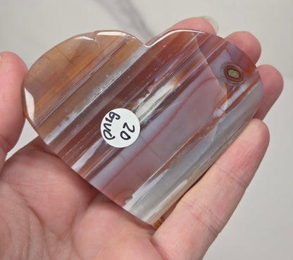 A hand holding a heart-shaped polished agate stone with horizontal banding in shades of brown, beige, and translucent white. A small white sticker with the handwritten text '20 BWD' is attached to the stone.