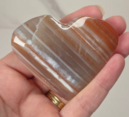 A hand holding a polished heart-shaped agate stone with natural banding in shades of brown, orange, and white. The person holding the stone wears a gold ring on their finger, and the background is a soft-focus light gray marble surface.