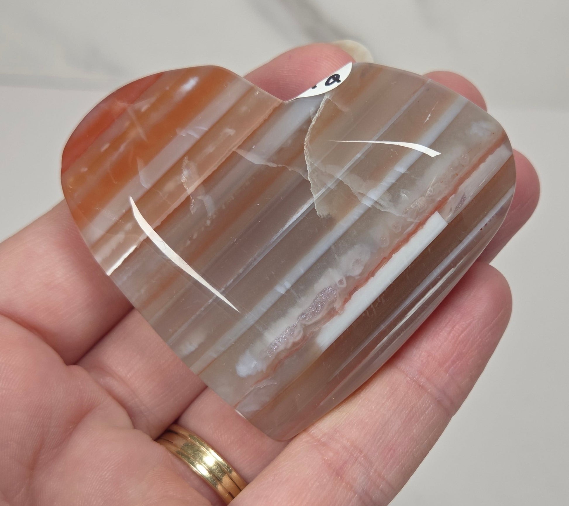 A heart-shaped polished agate crystal held in a person's hand. The stone features natural banding with colors of orange, brown, and translucent milky white, with a small white tag attached near the top.