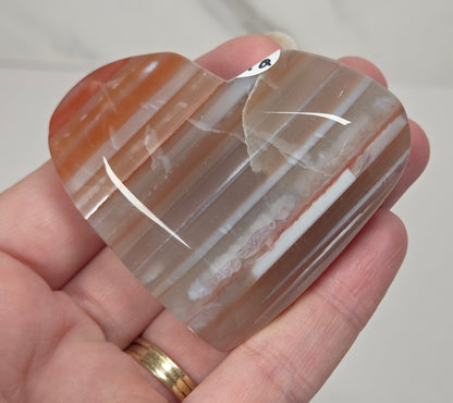 A heart-shaped polished agate crystal held in a person's hand. The stone features natural banding with colors of orange, brown, and translucent milky white, with a small white tag attached near the top.