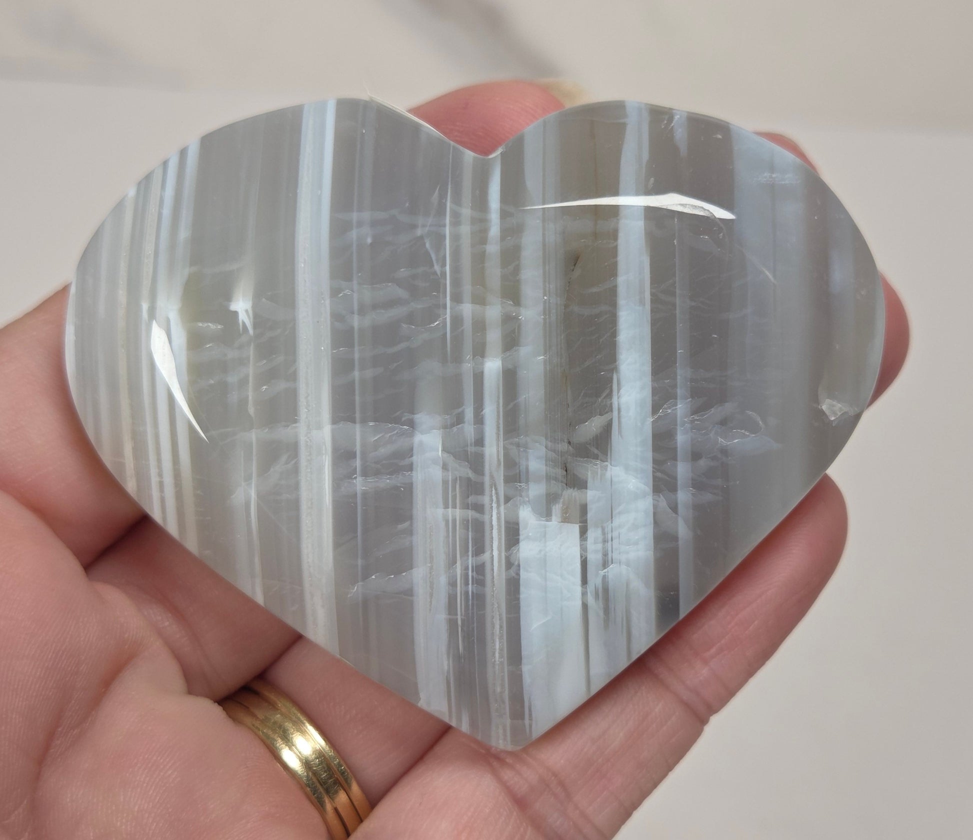 A heart-shaped polished stone with vertical white and gray banding, held in a person's hand against a light marble-like background.