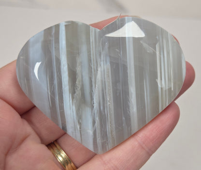 A heart-shaped polished stone with vertical striations in gray, white, and translucent tones, held in a person's hand against a light marble-like background.