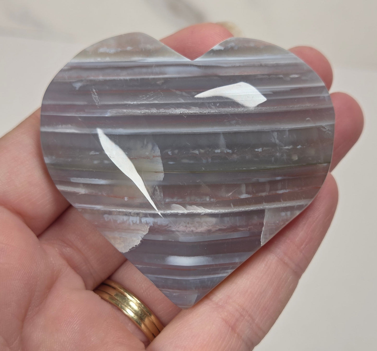 A heart-shaped polished stone with horizontal banding in gray, white, and muted purple tones, held in a person's hand against a light neutral background.