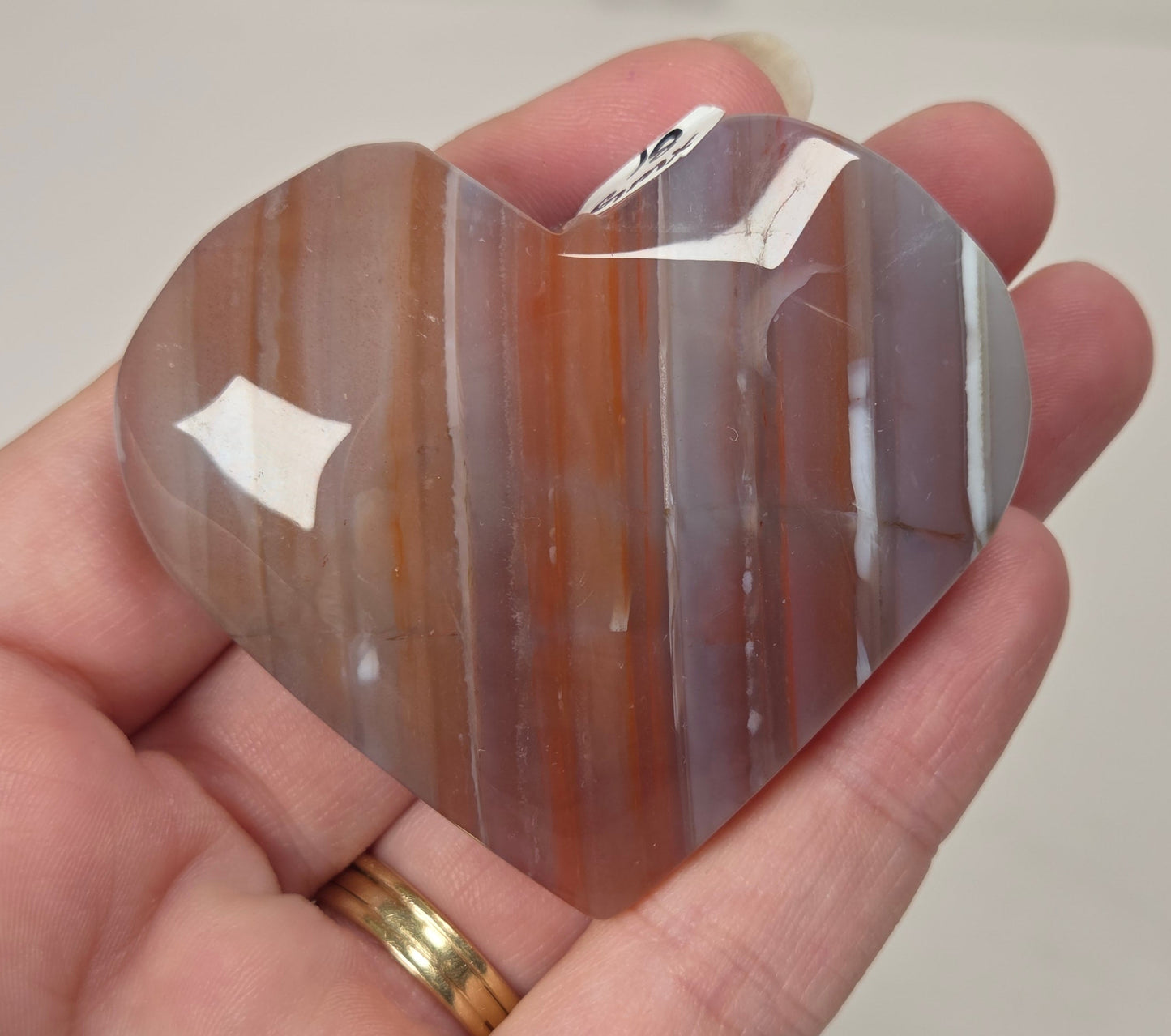 A heart-shaped polished stone with vertical bands of brown, orange, and gray, held in a person's hand. The stone has a small white tag attached near the top.