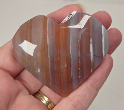 A heart-shaped polished stone with vertical bands of brown, orange, and gray, held in a person's hand. The stone has a small white tag attached near the top.
