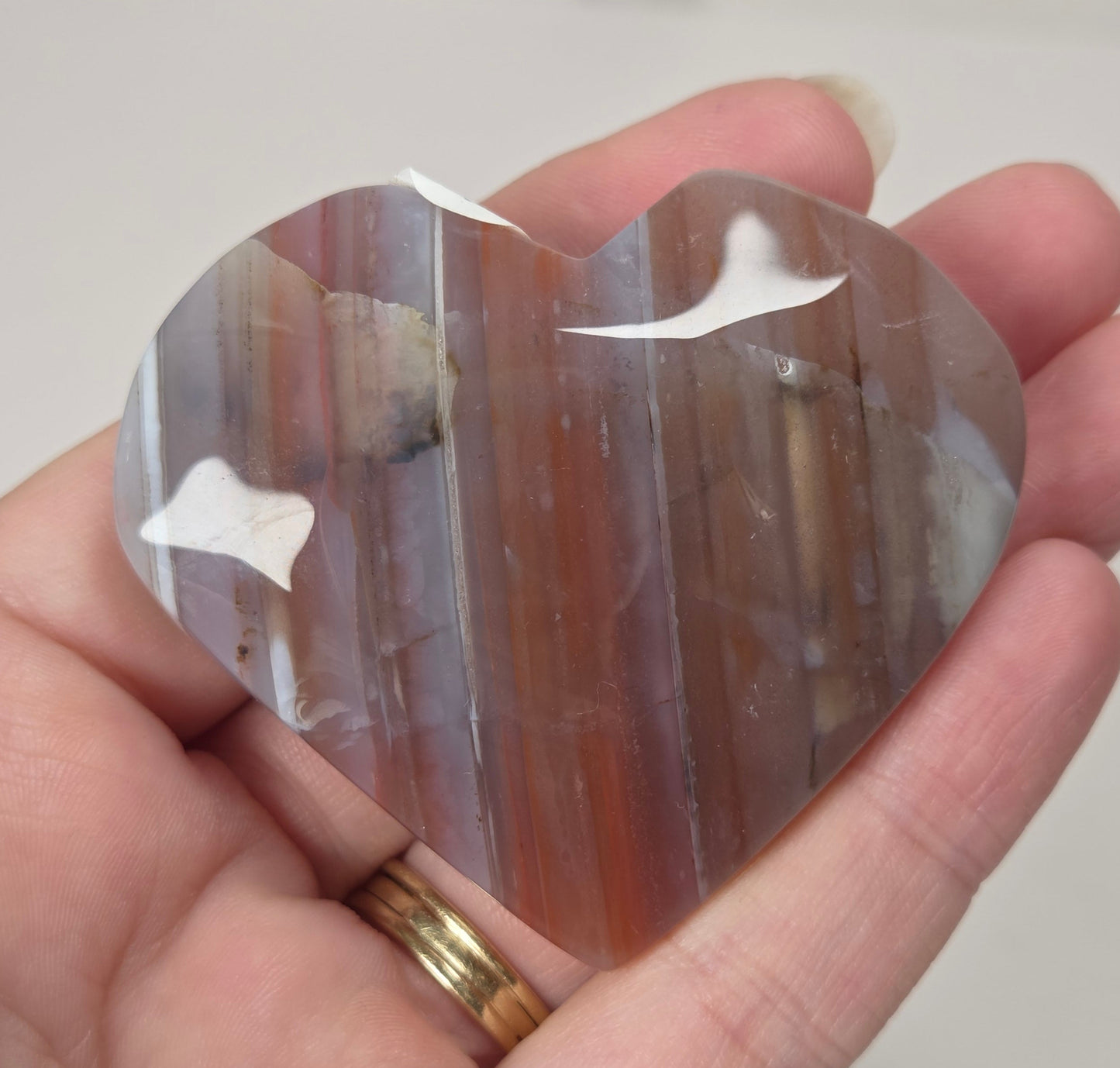 A heart-shaped polished agate crystal held in a person's hand. The stone features natural vertical banding with colors of brown, orange, and gray, and has white inclusions.