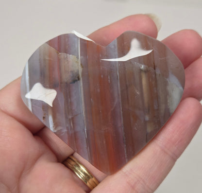 A heart-shaped polished agate crystal held in a person's hand. The stone features natural vertical banding with colors of brown, orange, and gray, and has white inclusions.