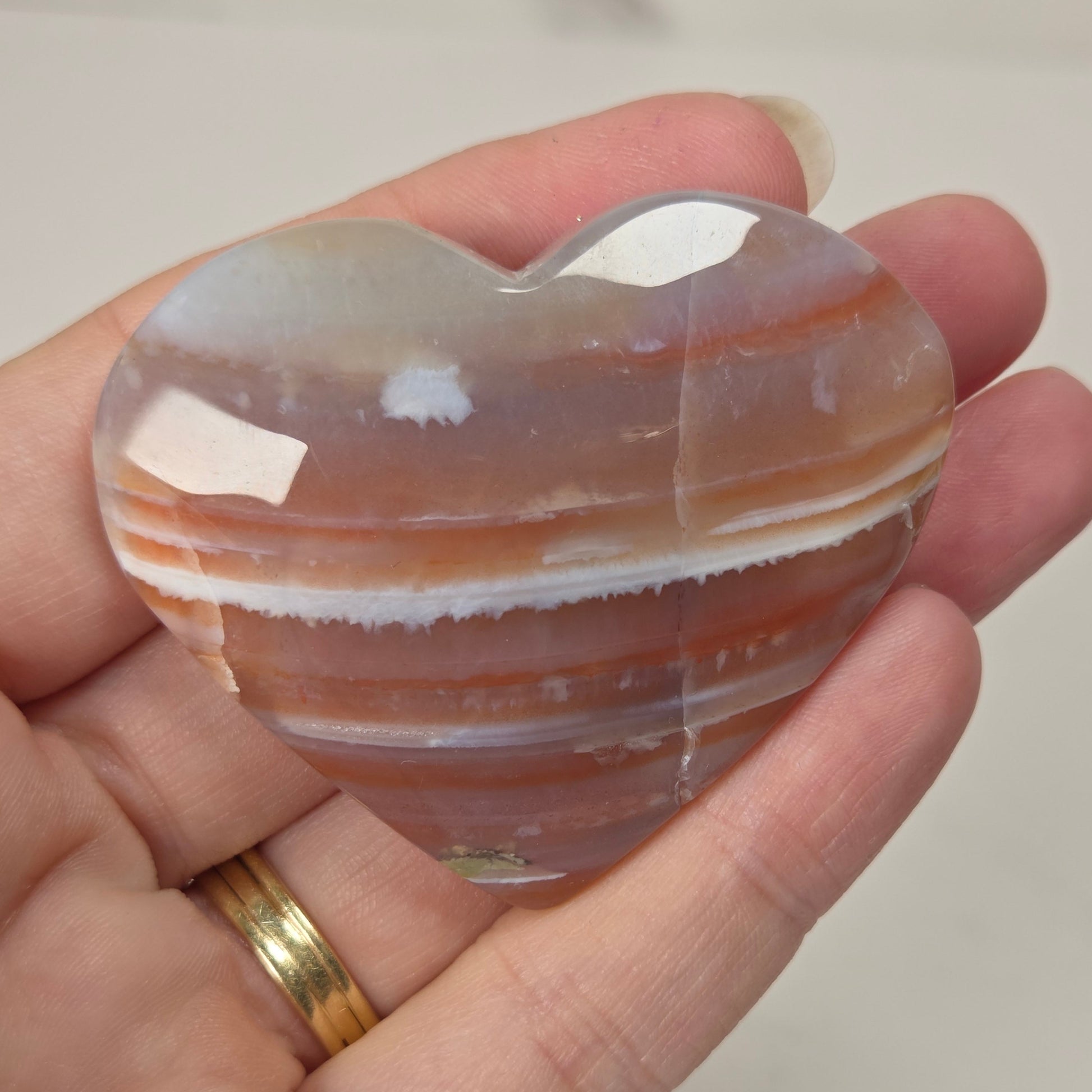 A heart-shaped polished agate crystal held in a person's hand. The stone features natural banding with colors of light gray, white, and warm reddish-brown.