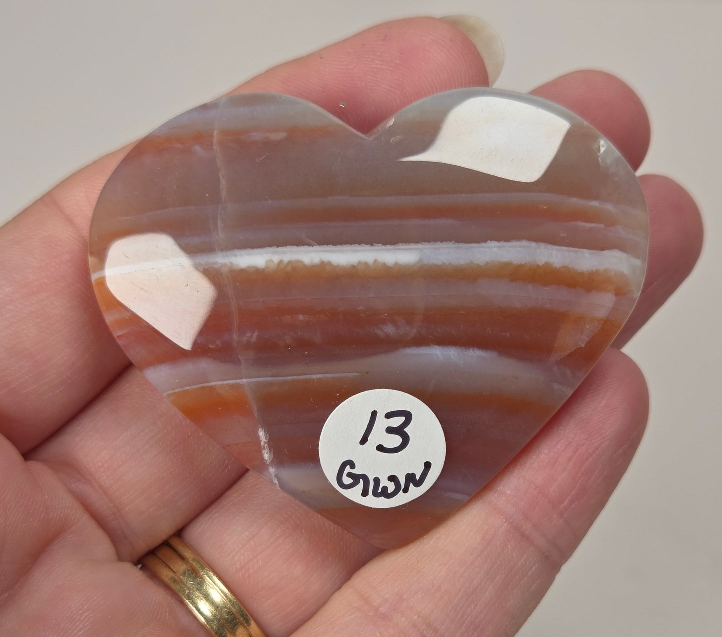 A heart-shaped polished agate stone with orange, brown, and white banded stripes, held in a person's hand. A small white sticker with the handwritten text '13 Green' is attached to the stone.